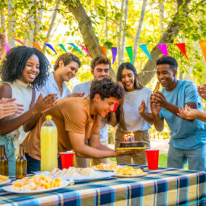 Young man celebrating birthday with friends at a park, blowing out candles on a cake while friends clap and cheer