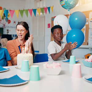 Birthday, cheering and children with teacher in classroom for group party with cake, hats and decoration. Happy, clapping hands and woman educator with kid students for celebration of special event.