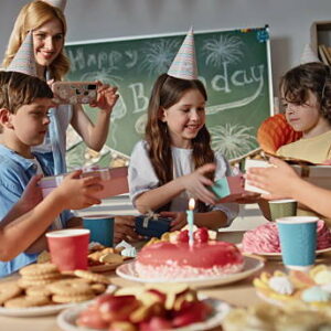 Kids celebrating birthday in classroom. Happy schoolgirl receiving gifts from group classmates. Joyful school children clapping hands congratulating smiling girl. Woman teacher shooting on cellphone