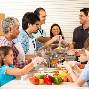 Multi-ethnic and mixed-age group of volunteers serve food to needy people at a community "soup kitchen" or food pantry location. The food has been donated by local neighbors. The volunteers wear aprons, lanyards, and gloves. Vegetables, fruits, and soup food items are on the table.  Service, kindness, charity, community outreach themes.